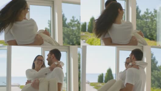 在阳光明媚的客厅里，一位英俊的年轻人举起他美丽的女友，他们旋转拥抱。他们是快乐的，房子是阳光和海滨风景。高清在线视频素材下载