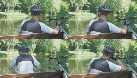 一个人骑着自行车，在公园里夏天休息。他坐在池塘边的长椅上。高清在线视频素材下载