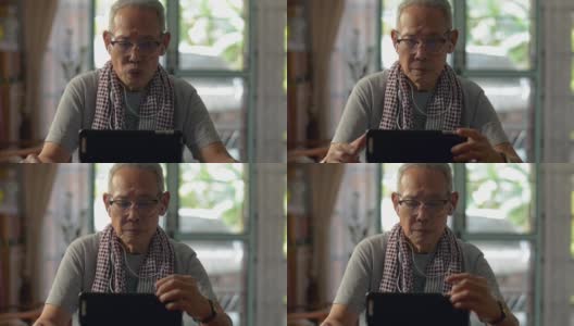 一位戴着眼镜、戴着耳机的亚洲老人在家里的桌子上用数码平板电脑。穿着休闲装的退休老人在数码设备上观看在线社交视频。回家隔离。老年人的生活方式。高清在线视频素材下载