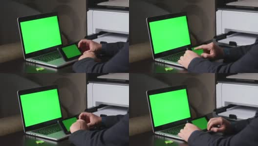 一名男子在一台绿色屏幕的笔记本电脑前工作。他手里拿着一个水平方向的绿色屏幕的智能手机。附近有一枚加密货币。Chromakey。高清在线视频素材下载