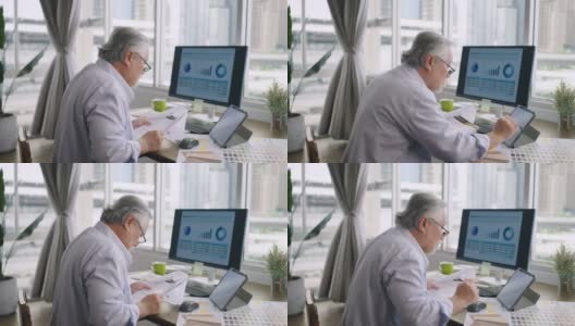 亚洲资深成熟男子在家里的桌子上用电脑和平板电脑工作，在忙碌中与客户交谈。高清在线视频素材下载