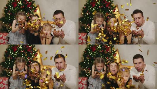 年轻幸福的四口之家坐在圣诞树旁，吹着金色的五彩纸屑。可爱的妈妈，爸爸和两个女儿一起庆祝圣诞节。Slowmotion拍摄高清在线视频素材下载