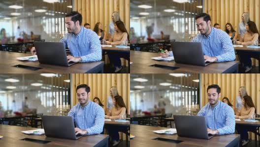 一个帅气的男人在一个共用的办公空间里用他的笔记本电脑工作，在他身后开会的一群人看起来都很高兴高清在线视频素材下载