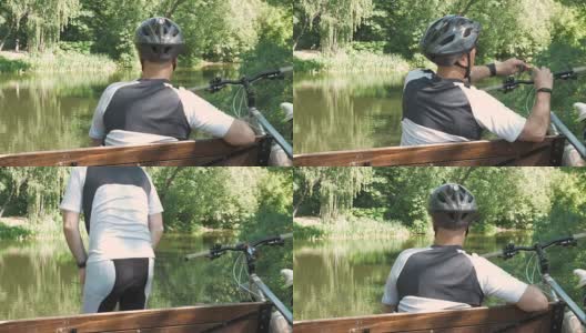 一个人骑着自行车，在公园里夏天休息。他坐在池塘边的长椅上。高清在线视频素材下载