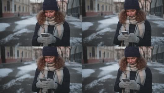 年轻女子戴着温暖的围巾和手套，微笑着看着屏幕上的智能手机输入信息在室外冬天的街道。高清在线视频素材下载