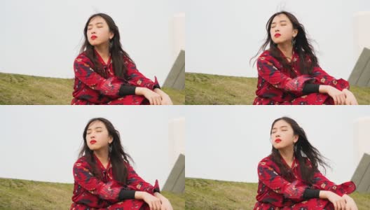 一个美丽的中国年轻女子坐在草地上，闭着眼睛，享受微风吹拂她的黑色长发，4k视频，慢镜头。高清在线视频素材下载