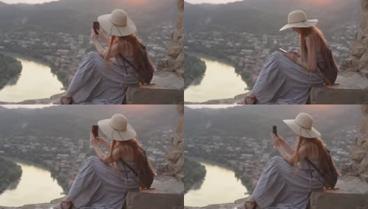戴着帽子背着背包的女孩坐在山上。拍摄视频到智能手机，城市下面高清在线视频素材下载