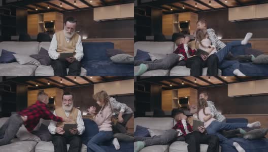 一个留着大胡子的80岁老人坐在沙发上用平板电脑工作，他的三个十几岁的快乐孙子跑过来拥抱他高清在线视频素材下载