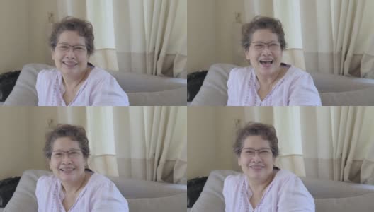 肖像幸福美丽的老年成人大龄中年亚洲女性戴着眼镜微笑大笑看着相机放松在客厅沙发上在家保健COVID-19疫情。高清在线视频素材下载