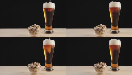 新鲜的啤酒。美味的精酿啤酒放在啤酒杯里，旁边放着一碟开心果，放在一张黑色背景的木桌上。冰凉的新鲜啤酒，带有水滴，泡沫和泡沫。广角镜头。4 k高清在线视频素材下载