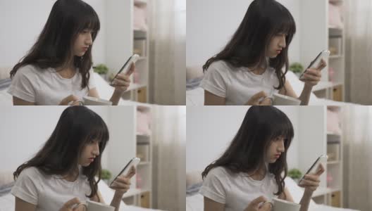 一名亚裔女孩在家里的卧室里端着茶杯，在智能手机屏幕上滚动着查看令人惊讶的网络新闻。高清在线视频素材下载
