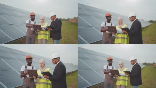 戴头巾的穆斯林妇女和戴白帽的印度男子与太阳能站的非裔美国技术人员交谈。多种族的人使用笔记本电脑，剪贴板和蓝图在户外工作高清在线视频素材下载