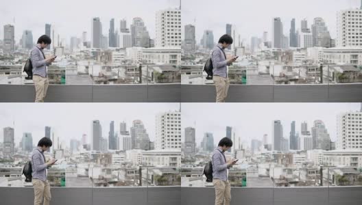 年轻的亚洲商人在办公大楼屋顶露台上使用智能手机，背景是城市景色。在智能设备上使用社交媒体应用程序或玩游戏。高清在线视频素材下载