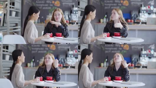 右图是两个年轻女性在咖啡馆一边喝咖啡一边愉快地聊天。红头发的女人情绪化地向她的亚洲朋友讲述故事高清在线视频素材下载