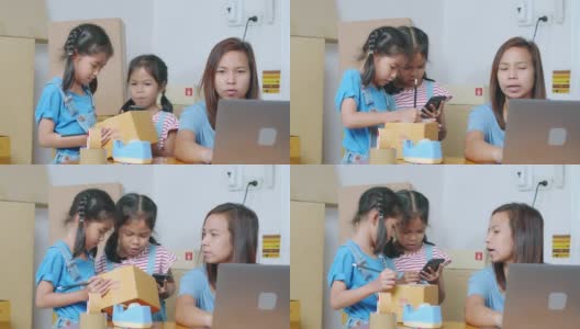 亚洲女孩正在把地址写进盒子里，以帮助父母为在线客户准备送货箱，而母亲使用笔记本电脑接收和检查网上购买的购物订单。高清在线视频素材下载