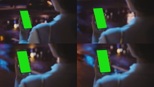 站着看绿屏的女人用Chroma Key智能手机霓虹夜市大街。使用手机的女性。过肩特写跟踪镜头聚焦显示高清在线视频素材下载