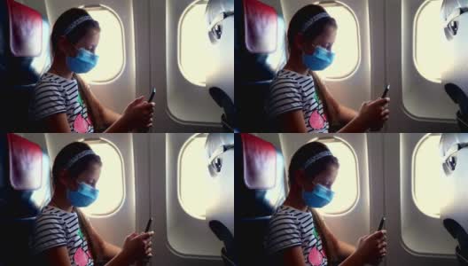 女孩、孩子戴着防护面具在飞机内使用手机、智能手机，坐在照明装置附近。新冠肺炎疫情后恢复航班。开放边界。Post-quarantine旅行高清在线视频素材下载