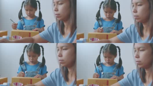 亚洲女孩正在把地址写进盒子里，以帮助父母为在线客户准备送货箱，而母亲使用笔记本电脑接收和检查网上购买的购物订单。高清在线视频素材下载