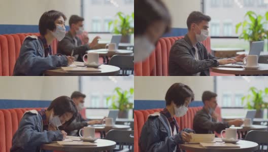 镜头从在笔记本电脑上工作的年轻白人男子，转向坐在咖啡馆桌旁浏览笔记本、做笔记和思考的亚洲女性，两人都戴着口罩高清在线视频素材下载