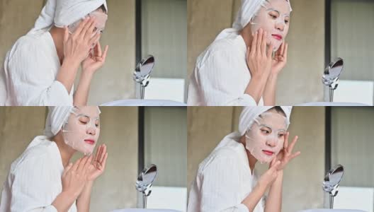 一段视频显示，一名亚洲女子一边对着镜子，一边敷面膜以改善皮肤。高清在线视频素材下载