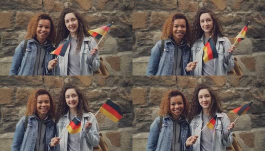 慢动作肖像两个微笑的女孩朋友挥舞着德国国旗，看着相机站在石头墙。友谊、旅游、快乐的人理念。高清在线视频素材下载