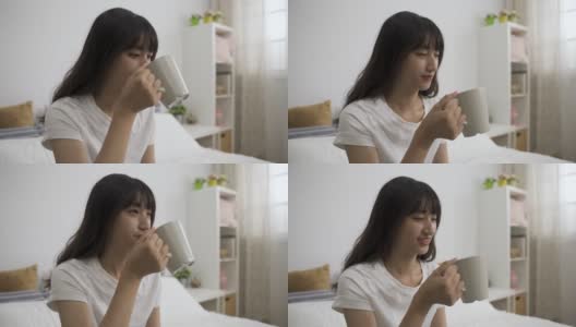阳光明媚的早晨，一位无忧无虑的韩国女性在家里的卧室里，面带满意地喝着美味的咖啡。高清在线视频素材下载