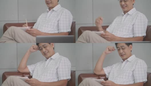 一位亚洲老人坐在房子的客厅里。带着快乐的微笑，在家里工作的技术概念高清在线视频素材下载