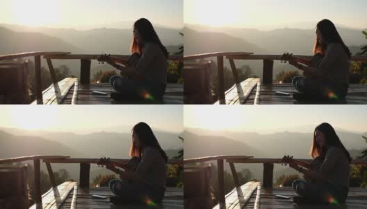 一个女性坐在和弹吉他的慢动作在一个美丽的山景日落前的木制阳台上高清在线视频素材下载