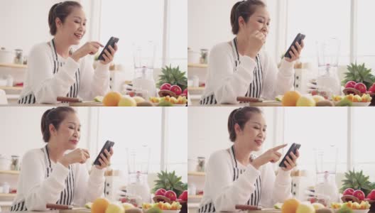 亚洲资深女性一边吃水果一边用手机感到幸福，准备用不同的口味和酸奶自制水果冰沙。健康天然益生菌调味饮料，在厨房学习使用科技高清在线视频素材下载