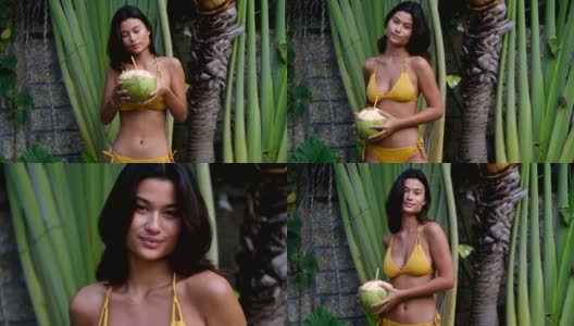 美丽的年轻亚洲女人在黄色比基尼在热带度假花园喝新鲜的年轻椰子。暑假高清在线视频素材下载