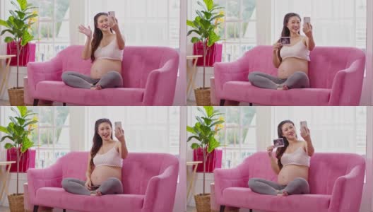快乐的孕妇坐在粉红色的沙发上，用手机和家人视频通话，在舒适的家里显示超声波图像。怀孕的年轻女子在家里享受着未来的生活高清在线视频素材下载