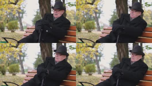 秋天的城市公园里，孤独的老爷爷拿着一根棍子坐在长椅上。高清在线视频素材下载