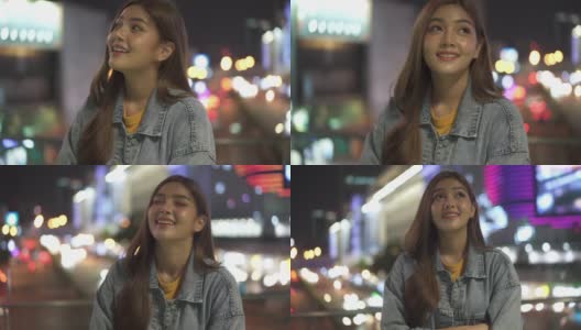 快乐年轻美丽的亚洲女人站在城市的夜晚，微笑着看着城市的灯光。漂亮女孩享受和享受在城市夜生活的乐趣。高清在线视频素材下载