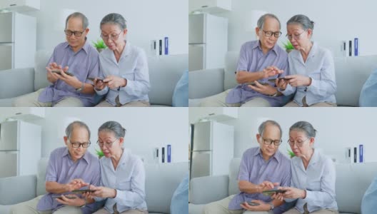 亚洲老年夫妇在家里的客厅里使用手机。强壮的老年人夫妇享受退休生活，在家里用智能手机一起看电影和看新闻。高清在线视频素材下载