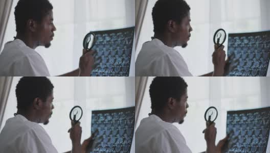 近距离聚焦的男医生站在窗口用放大镜分析x射线。侧视图肖像严肃、专注、聪明的非洲裔美国年轻人正在室内检查伦琴。高清在线视频素材下载