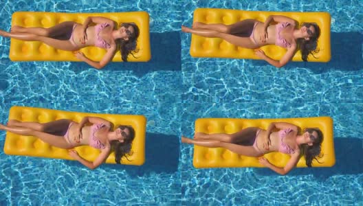 漂亮的女孩在太阳眼镜和比基尼躺在黄色的充气床垫在游泳池。在阳光明媚的日子里，年轻的女子在酒店的盆地里放松。暑假的概念。俯视图特写高清在线视频素材下载