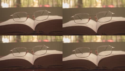 在一个温暖的阳光明媚的早晨，将一本打开的书和一副眼镜或眼镜放在木桌上，正面特写。孤立的背景。假日教育设计理念。高清在线视频素材下载