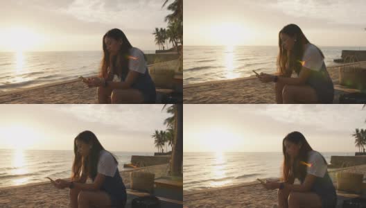 亚洲女人使用智能手机，坐在海边的海滩在早上日出的天空背景。独自快乐旅行的概念。4 k慢动作。高清在线视频素材下载