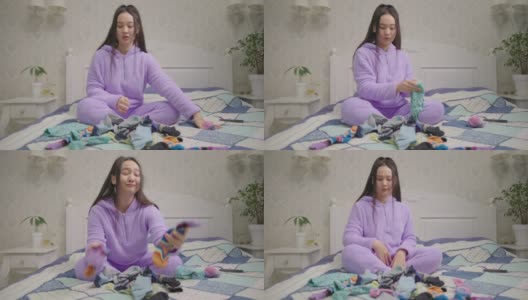 年轻的亚洲妇女折叠彩色袜子坐在床上。穿着紫色睡衣的女性通过整理衣服来整理和简化家庭。寻找袜子pair。高清在线视频素材下载