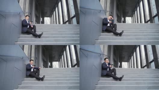 年轻的亚洲人沮丧地坐在楼梯上的街道附近的办公商业中心大楼高清在线视频素材下载