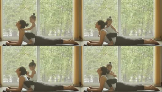 4 k。亚洲妈妈在家里练习瑜伽，有一个可爱的女儿坐在妈妈背上。隔离自我隔离期间的培训。休闲健康的生活方式。健康的家庭在早晨的活动高清在线视频素材下载