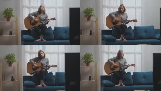 一个穿着灰色运动衫和黑色裤子的少女在吉他上弹奏着抒情的旋律。坐在蓝色沙发的背面。太阳从后面的窗户射进来。高清在线视频素材下载
