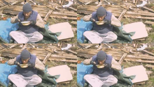 一个无家可归的人在废墟附近吃着盘子里的汤，在疫情期间帮助贫穷和饥饿的人们高清在线视频素材下载