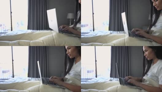 一个年轻的女人坐在房间的床上用笔记本电脑工作。工作、网上购物、查看电子邮件或在家里的床上搜索信息。高清在线视频素材下载