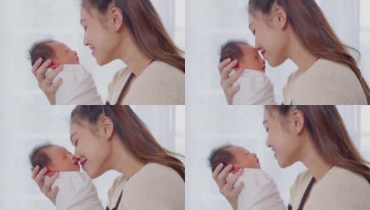 美丽迷人的亚洲妈妈抱着刚出生的宝宝，亲吻着宝宝的头，甜美可爱。快乐的妈妈和宝宝一起看着宝宝，带着爱微笑。母亲，婴儿，新生儿概念。慢动作高清在线视频素材下载