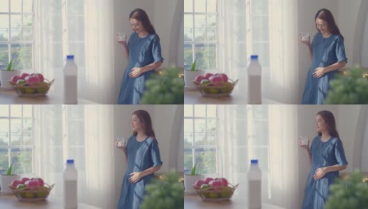 一位孕妇正在喝牛奶，看着窗户，轻轻地摸着她的肚子，怀孕/分娩高清在线视频素材下载