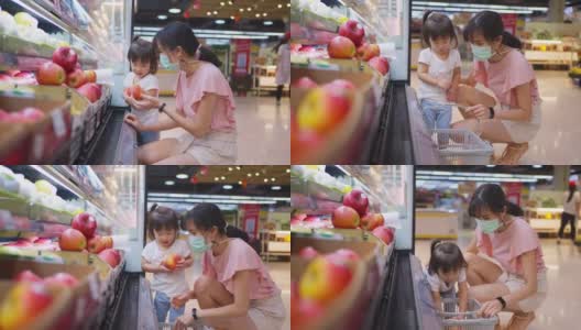 年轻美丽的亚洲母亲提着购物篮和她的孩子在超市行走，戴着口罩，以预防新冠肺炎感染。她正在挑选红苹果水果并从货架上挑选。高清在线视频素材下载