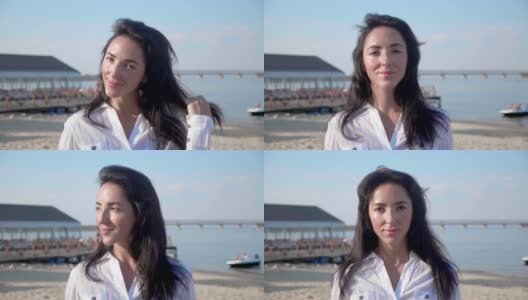 年轻女子的肖像看着相机站在海滩上与大海的背景。穿着白色t恤的漂亮女孩抬起了眼睛。风吹拂着轻盈的棕色头发，在阳光下闪闪发光高清在线视频素材下载