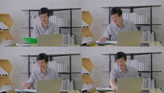 一名亚洲高级男子正坐在一间办公室里，看着一台笔记本电脑，面带微笑地工作着，笑容满面，事业成功，在家工作高清在线视频素材下载
