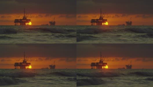 慢动作拍摄的太阳在地平线上落下，海浪拍打着海岸的亨廷顿海滩在南加州与几个近海石油钻井平台和石油(石油)油轮的剪影在戏剧下的远方高清在线视频素材下载